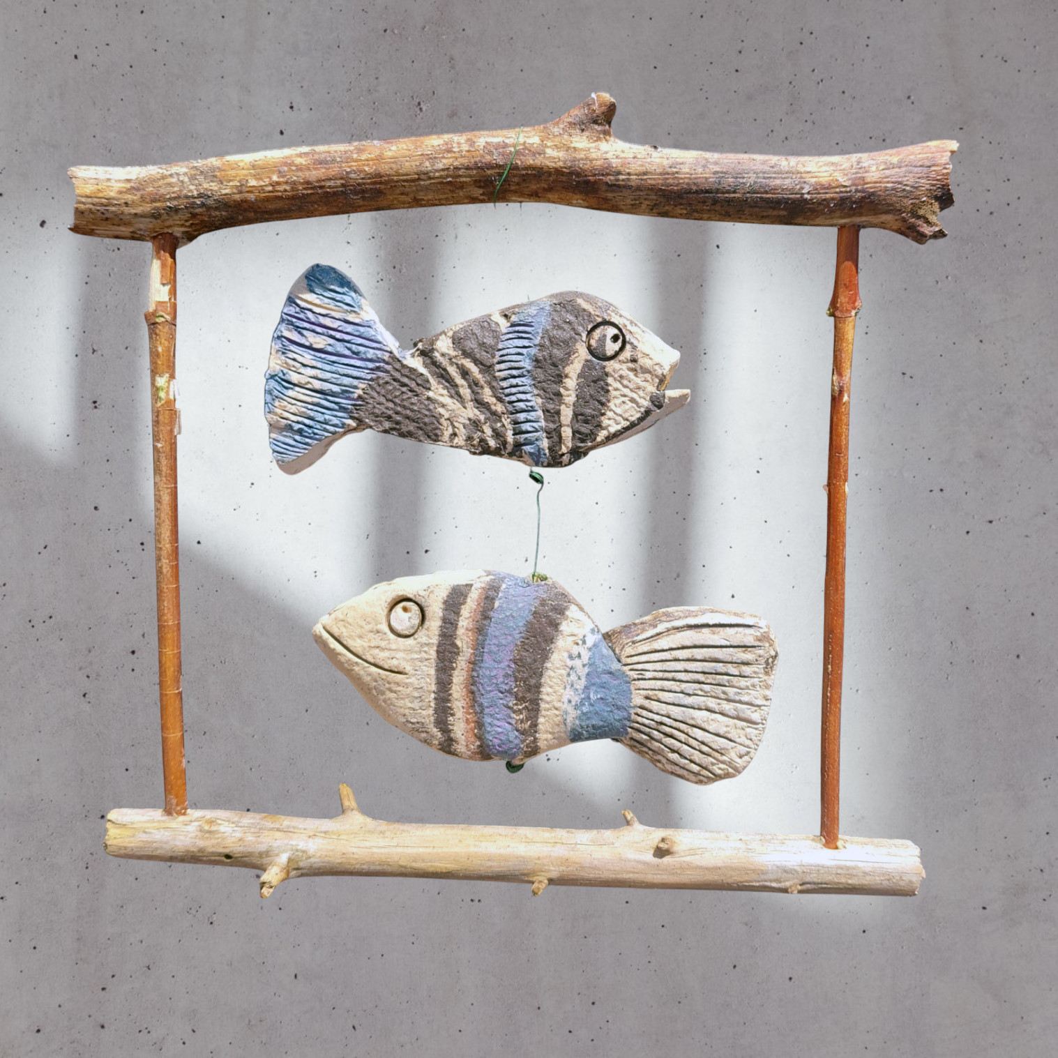 Zwei handgefertigte Keramikfische in Blau- und Erdtönen, hängend an einem Holzrahmen, ideal für maritime Fensterdekoration.