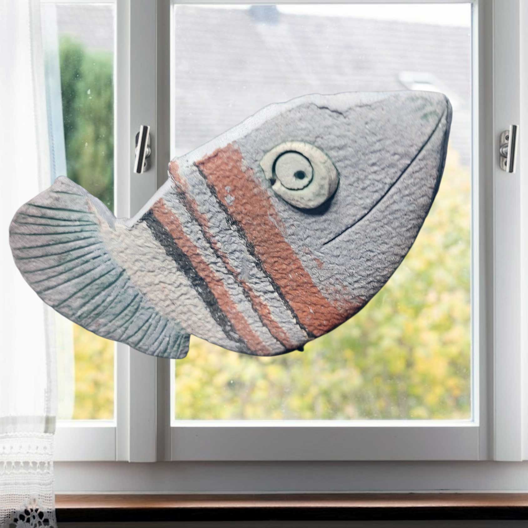 Ein handgefertigter, gestreifter Keramik-Fisch als maritime Fensterdekoration mit strukturierter Oberfläche vor einem hellen Fenster im Innenraum.