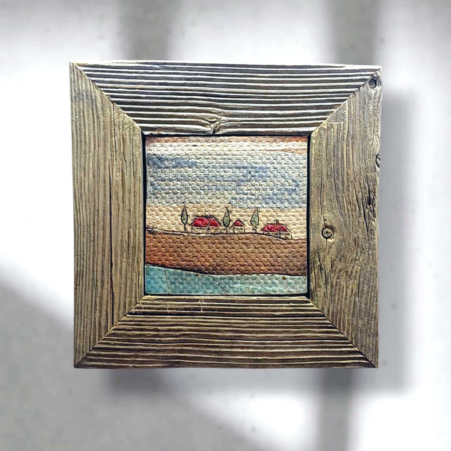 Handgearbeitete Keramikfliese mit naiver Landschaftsdarstellung in einem rustikalen Rahmen aus Altholz, ideal als dekoratives Wandbild für das Zuhause.